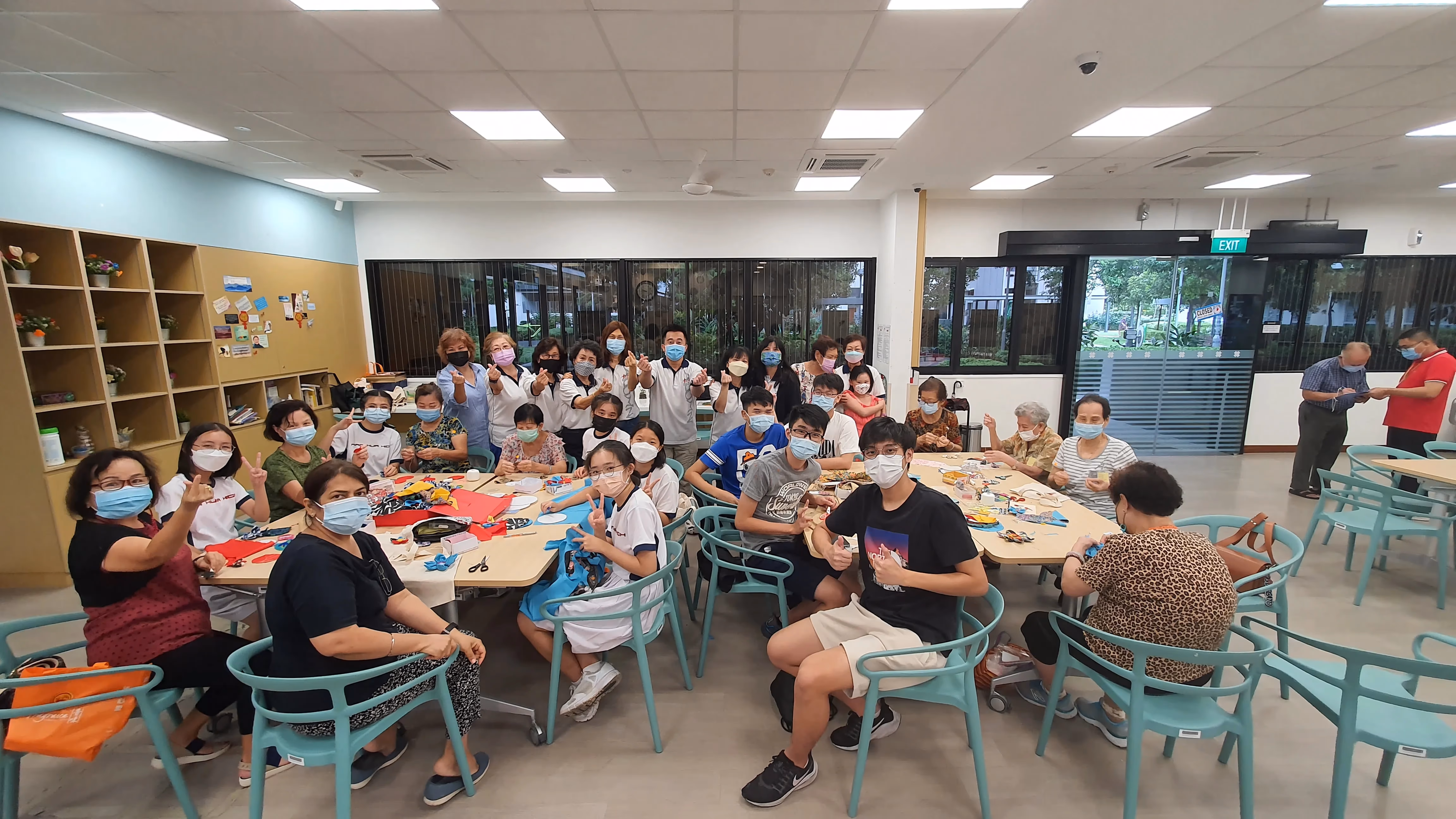 A large group of masked people of various ages sit around tables doing crafts in a bright community room.